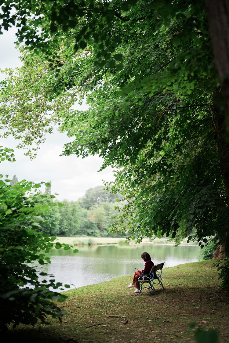 A woman resting on a blue iron bench at the water's edge, tucked under tall trees in the park at Ermenonville, France.