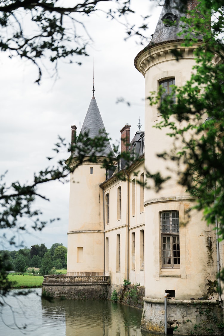 Twin turrets of Domaine du Château d'Ermenonville rising over the moat, framed through dark oak branches on the bank.