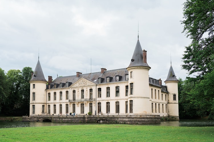 The four-turreted facade of Domaine du Château d'Ermenonville standing over its moat and wide green lawn beneath a soft cloudy sky.