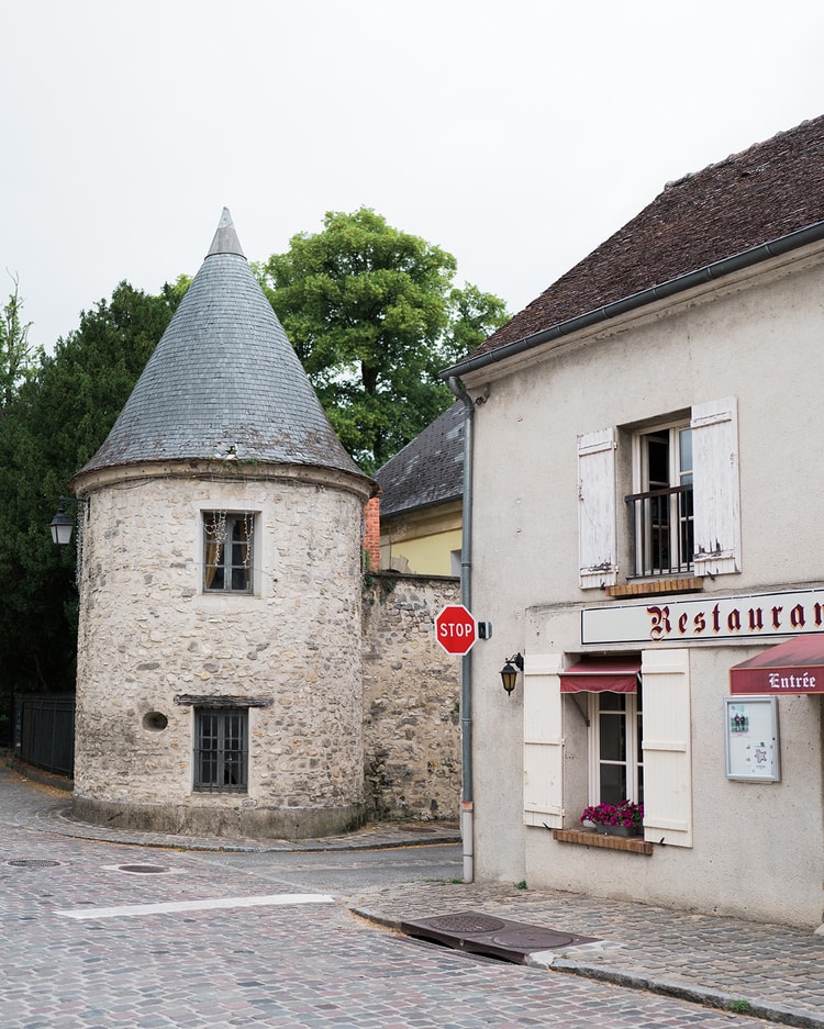 Round stone turret with a pointed slate roof beside a shuttered restaurant in Ermenonville, France, a red stop sign marking the cobbled corner.