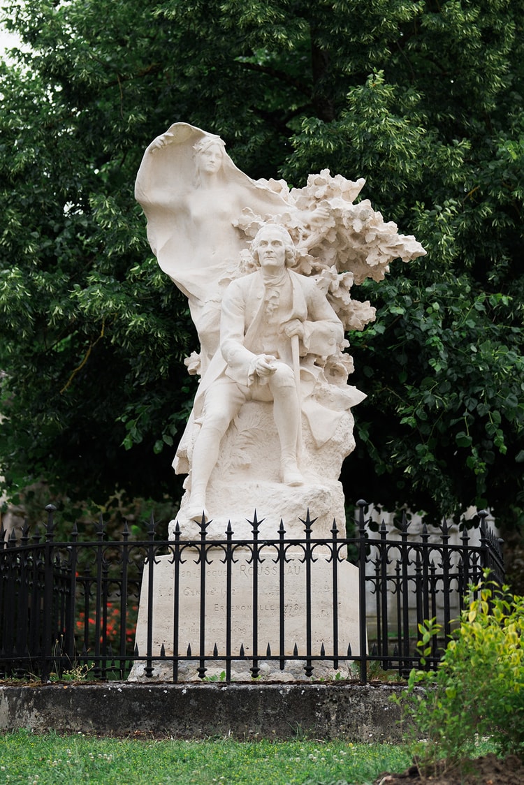 Stone monument to Jean-Jacques Rousseau in Ermenonville, France, the seated philosopher backed by a veiled allegorical figure behind a black iron fence.