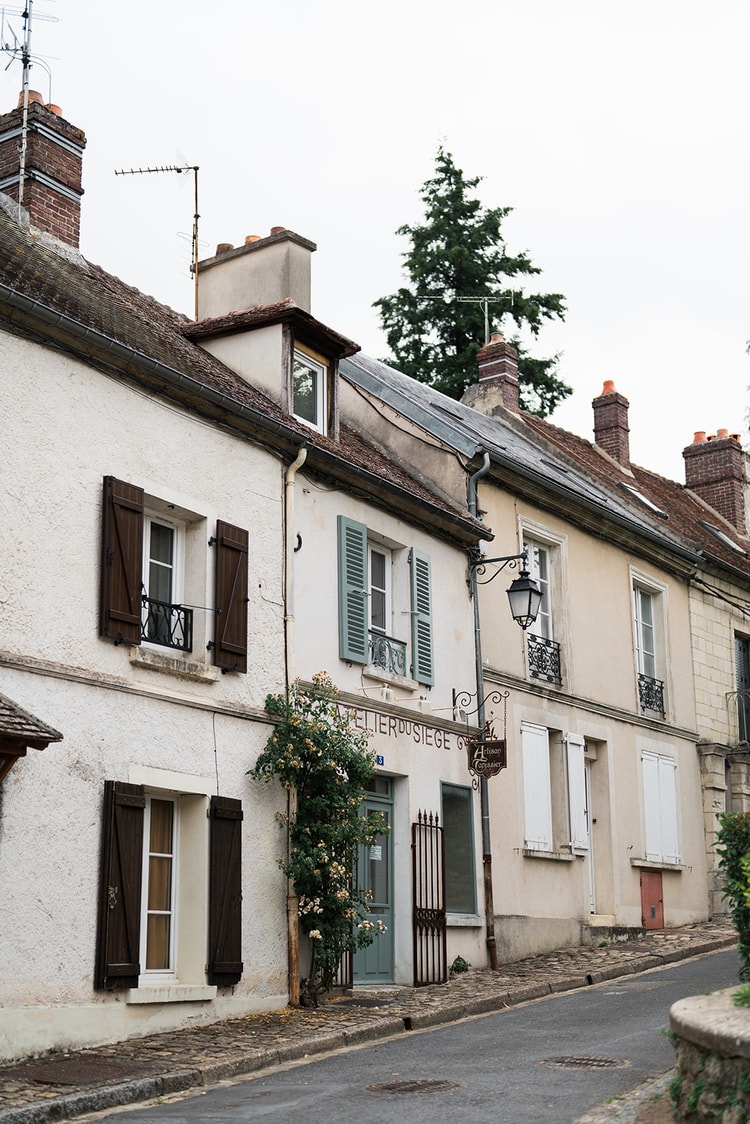 Row of shuttered houses on a sloping street in Ermenonville, France, a climbing rose framing the doorway of an upholsterer's atelier.