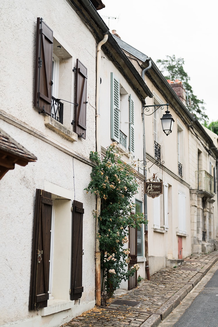 Shuttered houses along a cobbled street in Ermenonville, France, with an iron lantern and a wrought-iron Artisan Tapissier sign above a climbing rose.