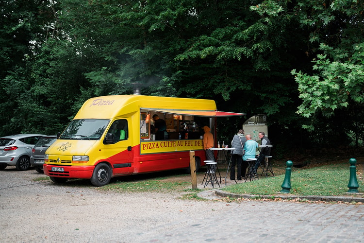 Smoke rising from a wood-fired pizza truck near the park entrance in Ermenonville, France, as locals order at the window under the trees.