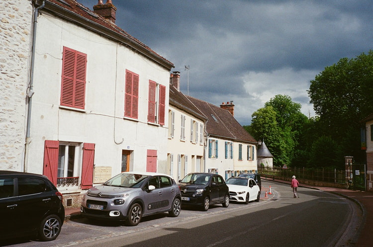 Red-shuttered houses in Ermenonville, France, catching low sun against a dark storm sky, cars parked along the curving street.