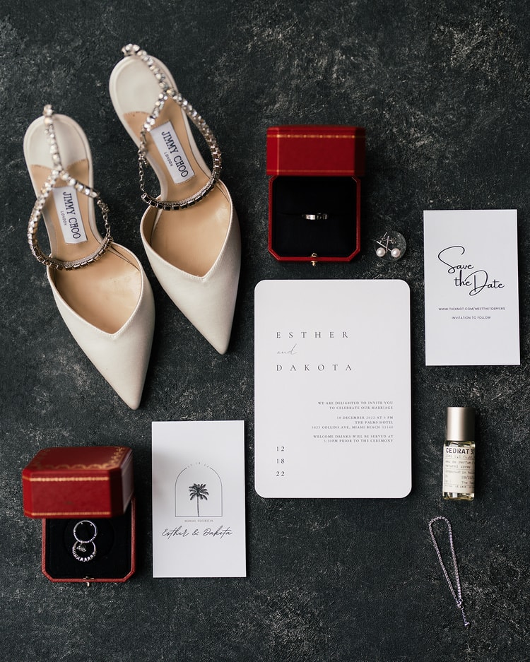 Bridal detail flat lay on dark stone: pearl-strap Jimmy Choo heels, two red ring boxes with wedding bands, invitation suite, Save the Date card, pearl stud earrings, and a perfume bottle
