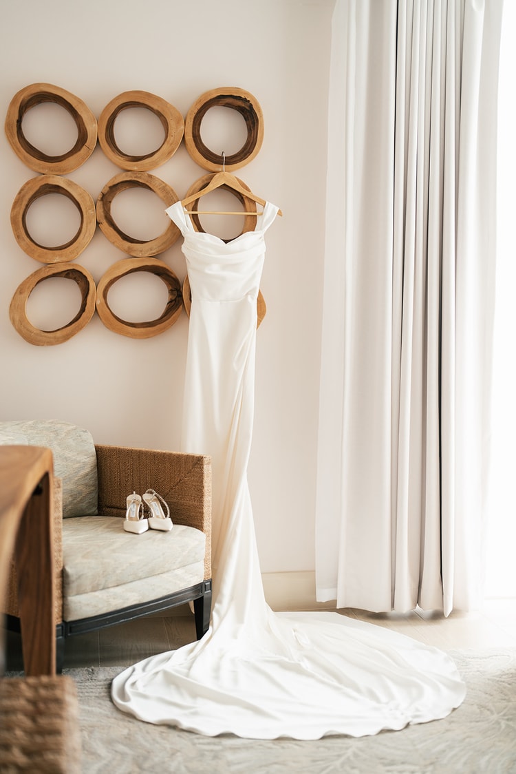 White off-shoulder gown on a hanger against a circular wood-ring wall sculpture beside a wicker chair, bridal shoes resting on the seat, wedding details
