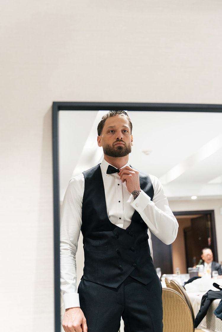 Groom in a black tuxedo vest and bow tie adjusting his collar in front of a large mirror, wedding photography