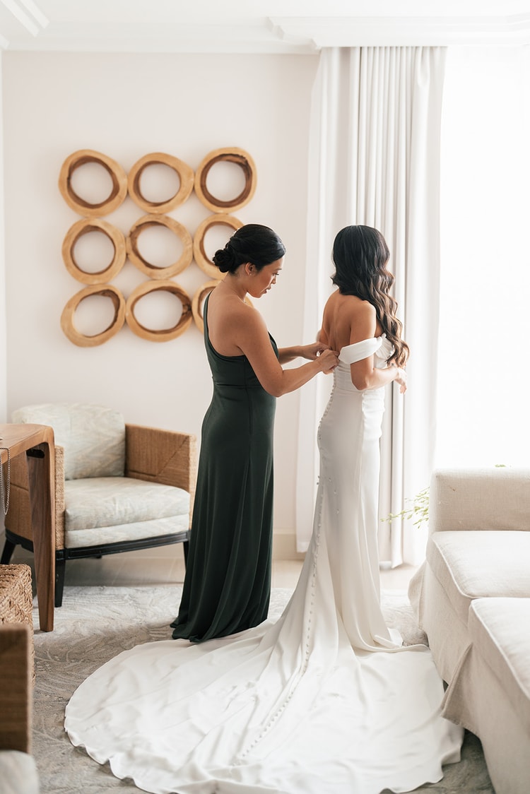 Bridesmaid in a forest green gown fastening the back of the bride's white off-shoulder gown in a bright hotel suite, wedding photography