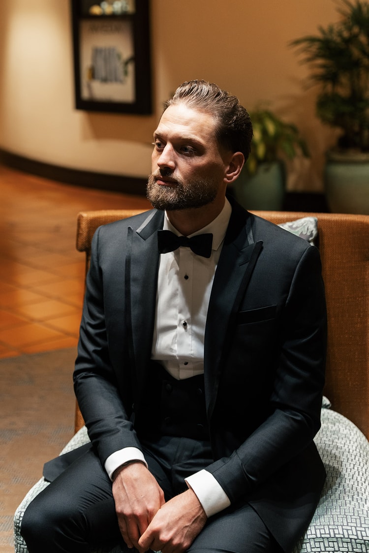 Groom in a black tuxedo and bow tie seated in a plush chair, gazing to the side in a warmly lit corridor, wedding photography