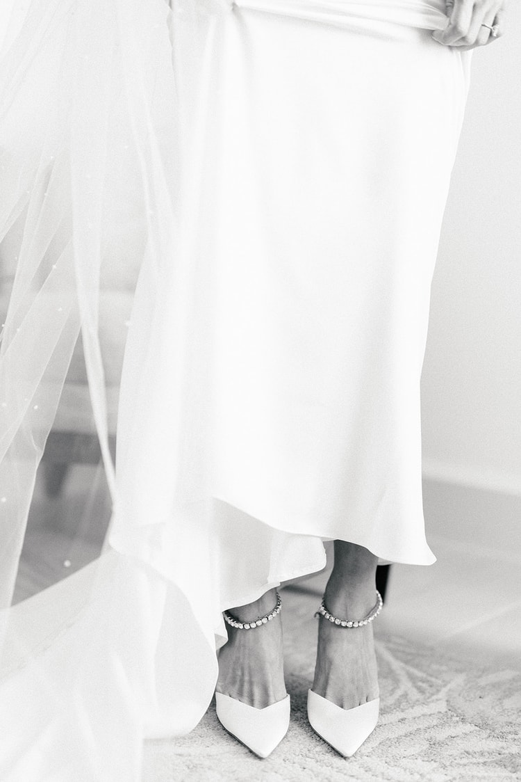 Close-up of bride's feet in crystal-strap pointed-toe heels beneath a flowing white gown and pearl-dotted veil, wedding details