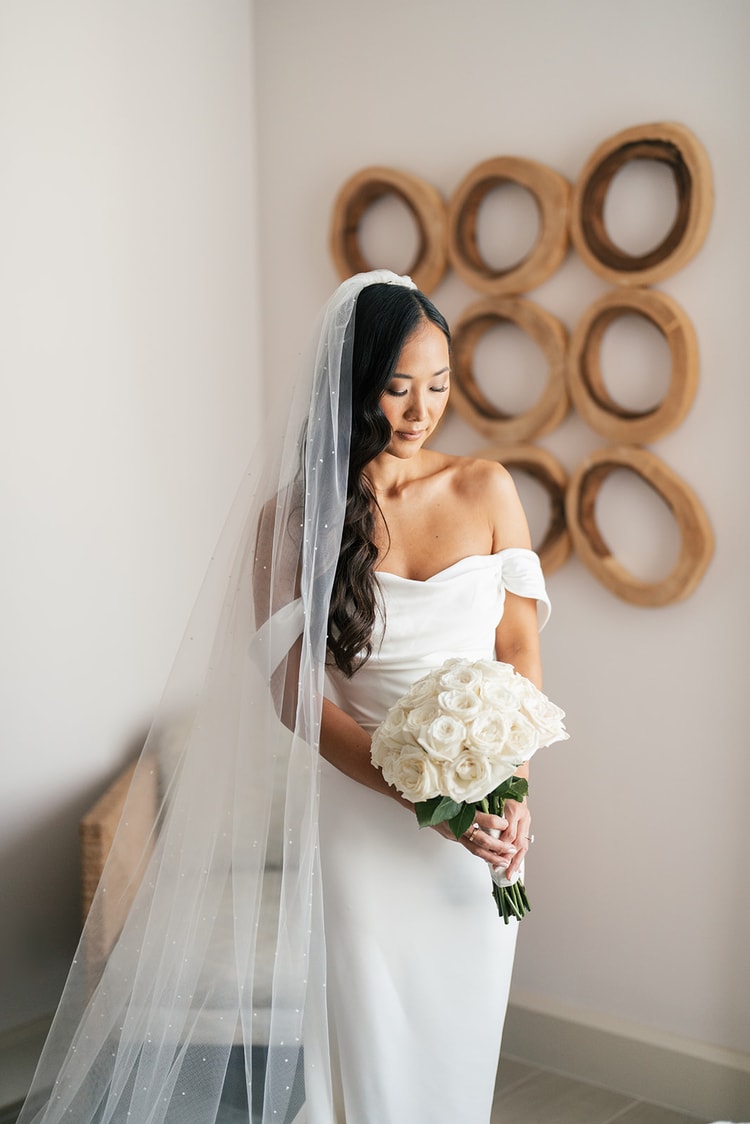 Bride in an off-shoulder white gown with a pearl-dotted cathedral veil holding a tight white rose bouquet, gazing down in a bright room, wedding photography