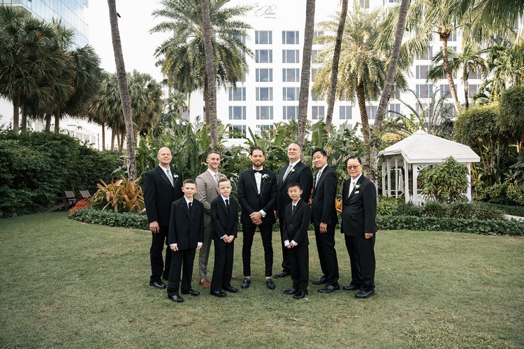 Groom with groomsmen and young boys all in dark suits posing on a lawn with tall palm trees and white hotel building behind, wedding photography