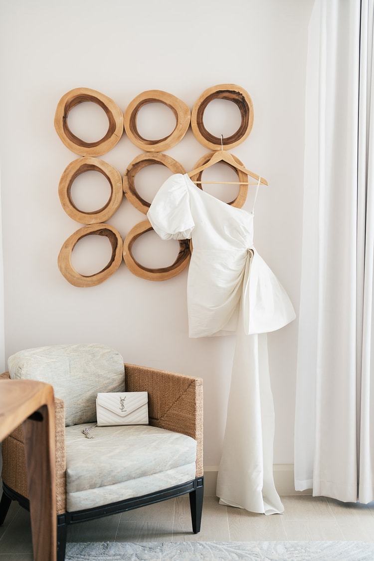 White one-shoulder mini dress with a puffed sleeve and draped skirt on a hanger against a circular wood-ring wall sculpture, a white YSL clutch resting on a rattan armchair below