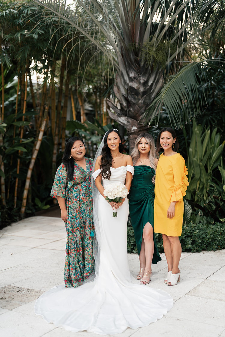 Bride with three guests in colorful dresses, green and yellow, posing together before a backdrop of palm trees, wedding photography