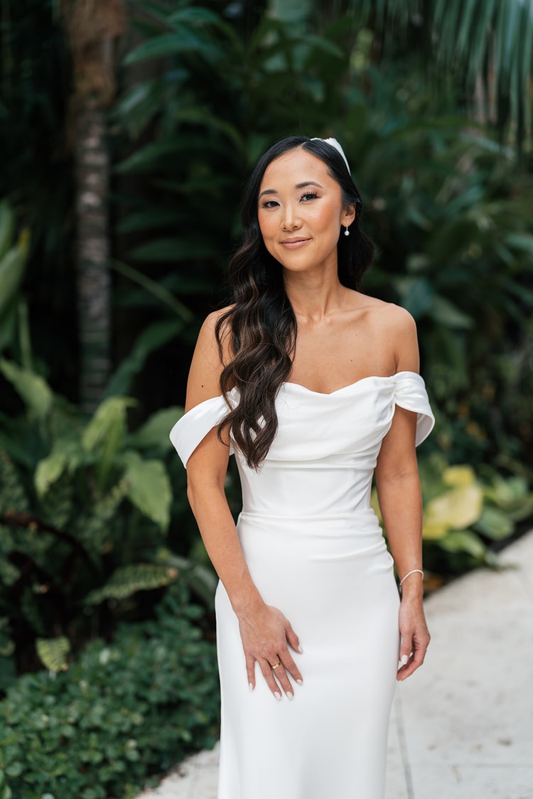 Bride in an off-shoulder gown smiling at the camera, surrounded by dense tropical greenery, wedding photography