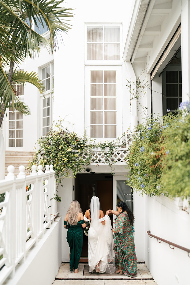 Bride and two bridesmaids in deep green dresses entering a white building draped with flowering vines, wedding photography