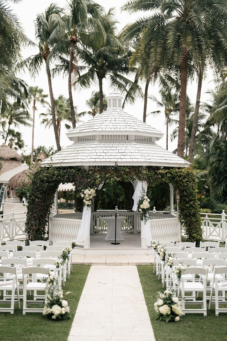 Outdoor ceremony space with a white ivy-covered gazebo, white folding chairs on a lawn decorated with white floral arrangements, palm trees overhead, wedding photography
