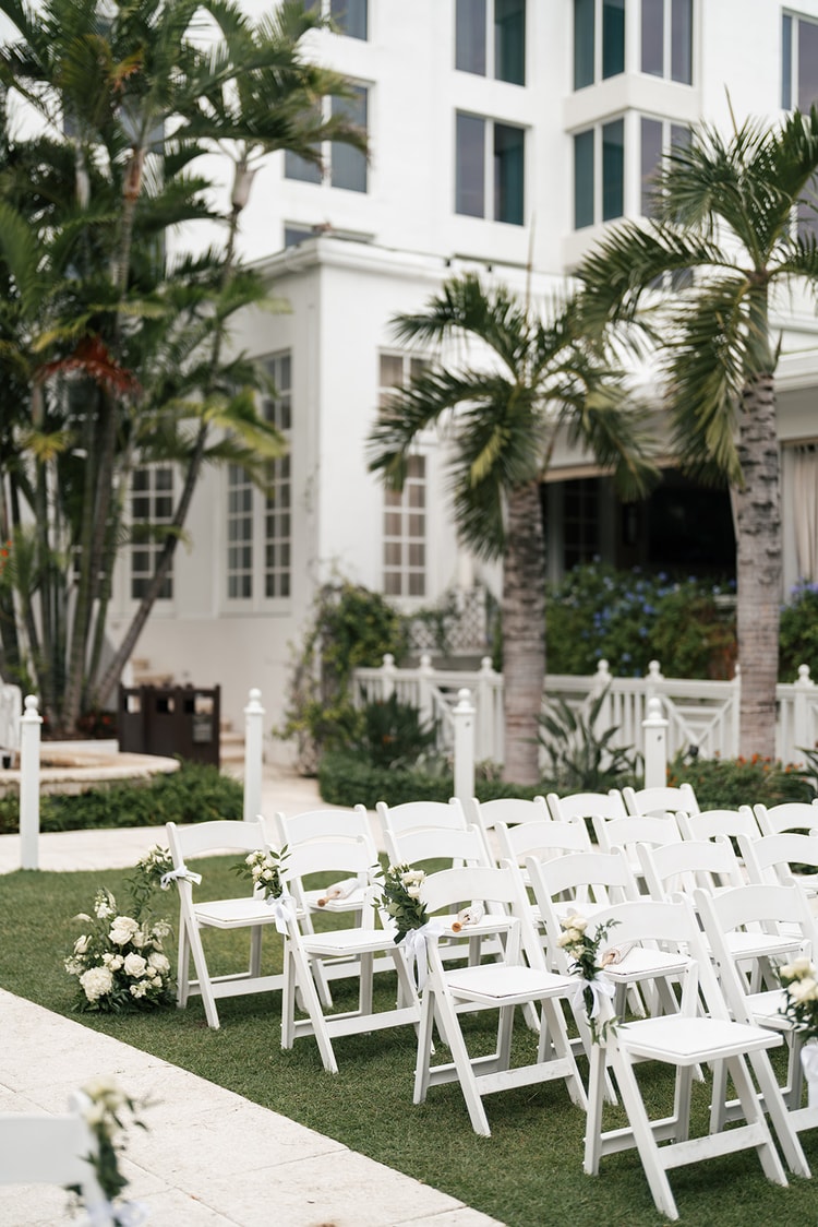 White folding chairs lined up on a green lawn with white floral chair accents, a white colonial building and palm trees in the background, wedding photography