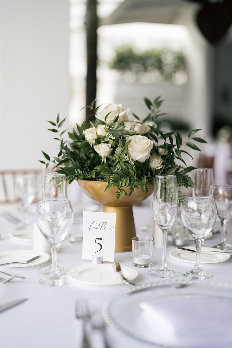 Reception centerpiece: white roses and ferns in a golden pedestal bowl, table number card and crystal glassware on a white linen table, wedding details