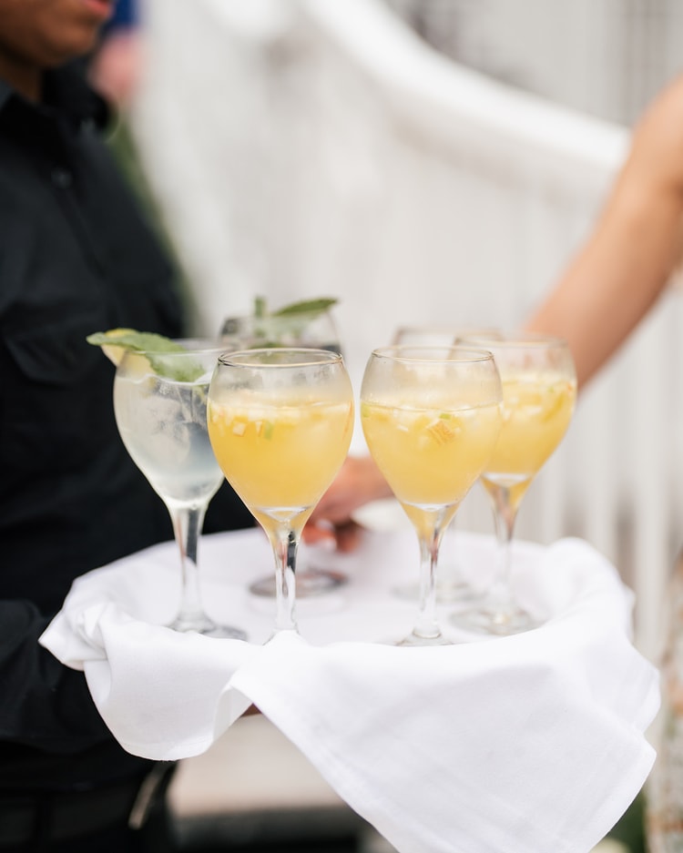 Server holding a white-draped tray of yellow and clear cocktails garnished with fresh herbs at an outdoor reception, wedding photography