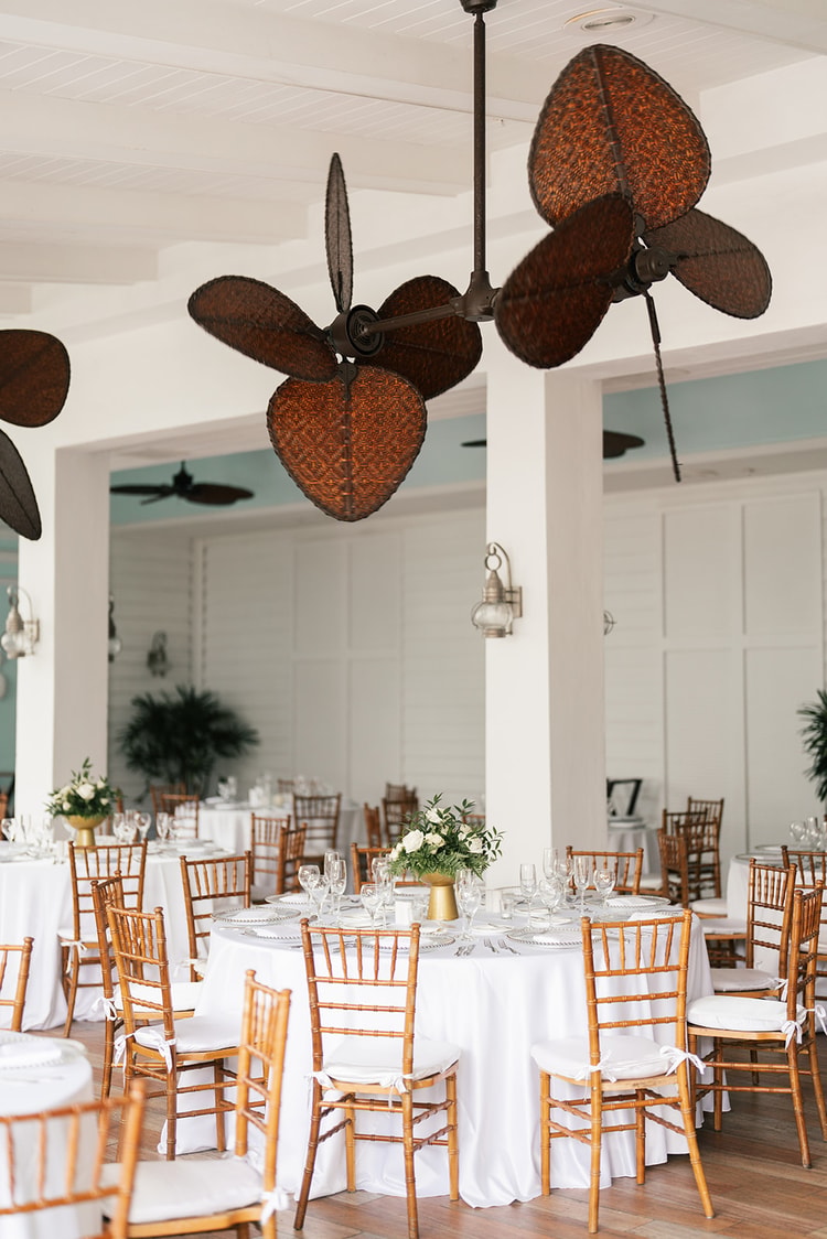 Open-air reception ballroom with white linen round tables, bamboo Chiavari chairs, gold vase centerpieces with white florals, and large ornate wicker ceiling fans overhead