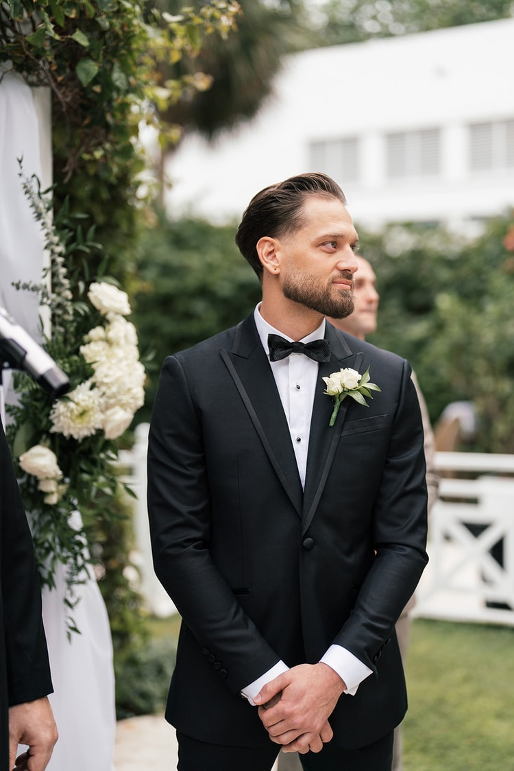 Groom in a black tuxedo with a white rose boutonniere waiting at the altar, watching down the aisle with a soft smile, wedding photography
