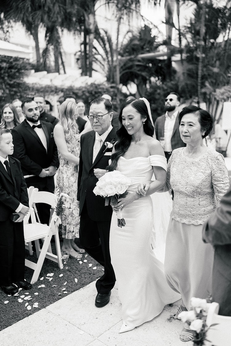 Bride walking the aisle escorted by her parents, smiling, guests watching from white folding chairs, palm trees and lush greenery behind, black and white, wedding photography