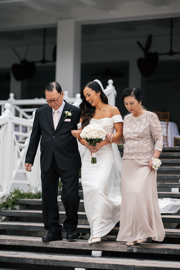 Bride laughing as she descends broad wooden steps escorted by both parents, white rose bouquet in hand, wedding photography