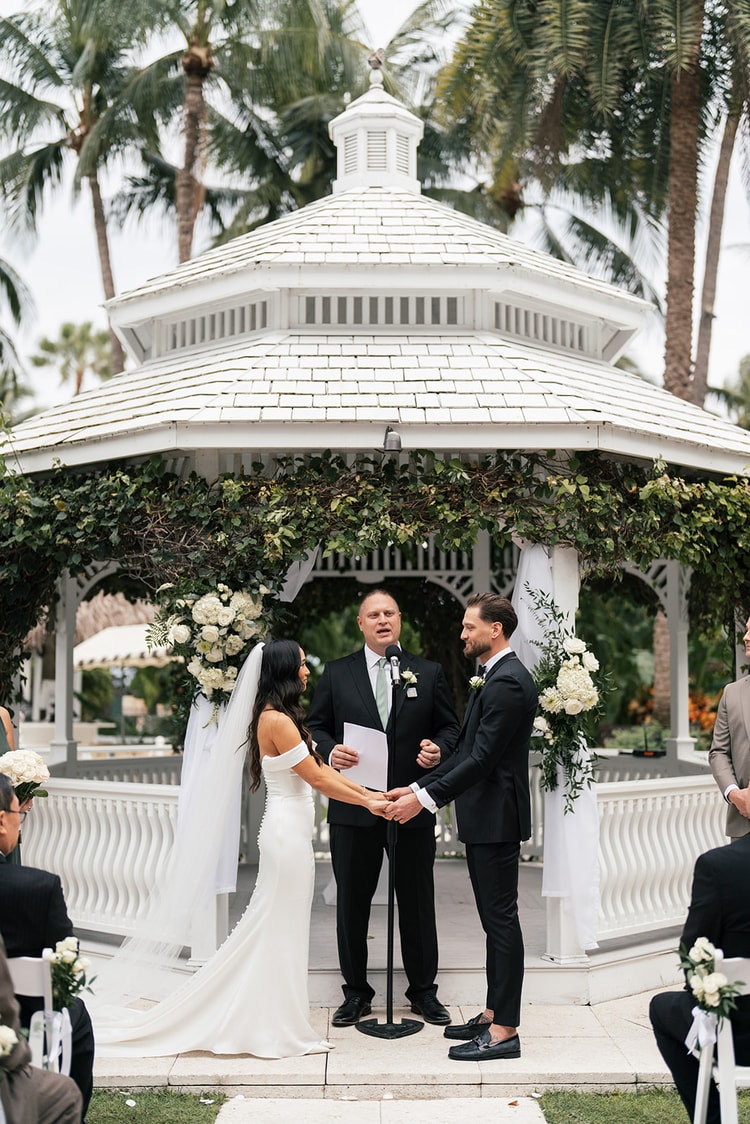 Couple exchanging vows beneath an ivy-draped white gazebo surrounded by tall palm trees, guests seated on the lawn, wedding photography