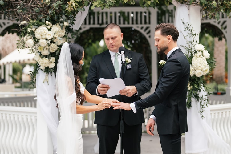 Couple exchanging rings at a white gazebo altar adorned with white floral arrangements and lush greenery, officiant reading from papers, wedding photography