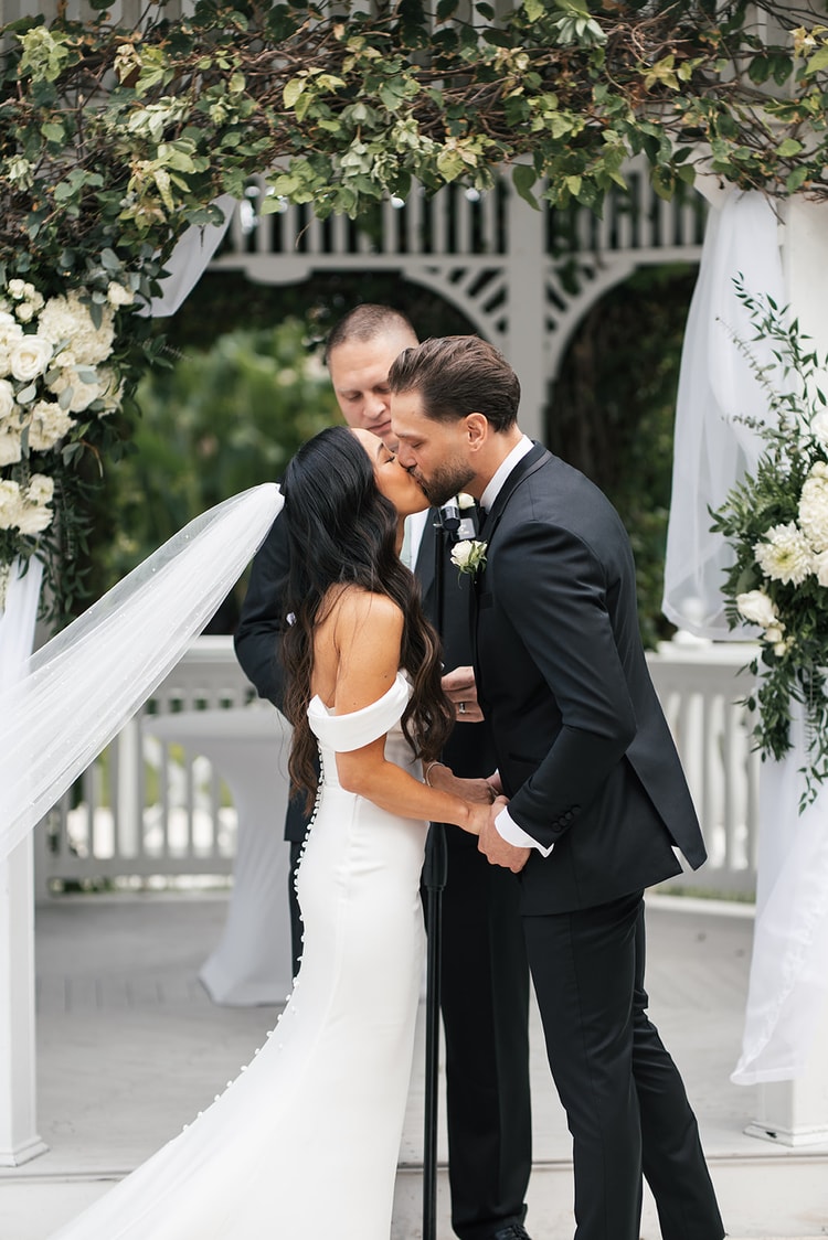 Bride and groom share their first kiss beneath a white gazebo framed by white florals and greenery, officiant standing behind, wedding photography