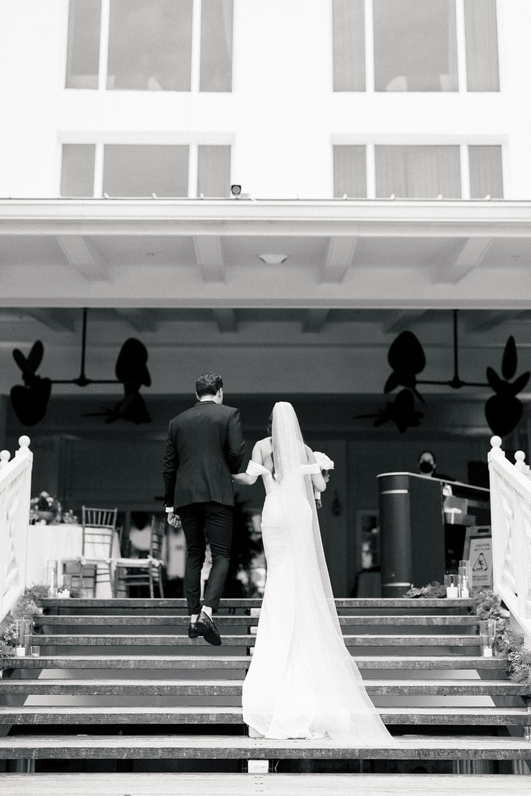 Newlywed couple walking hand in hand up broad outdoor stairs toward a covered venue, seen from behind in black and white, wedding photography