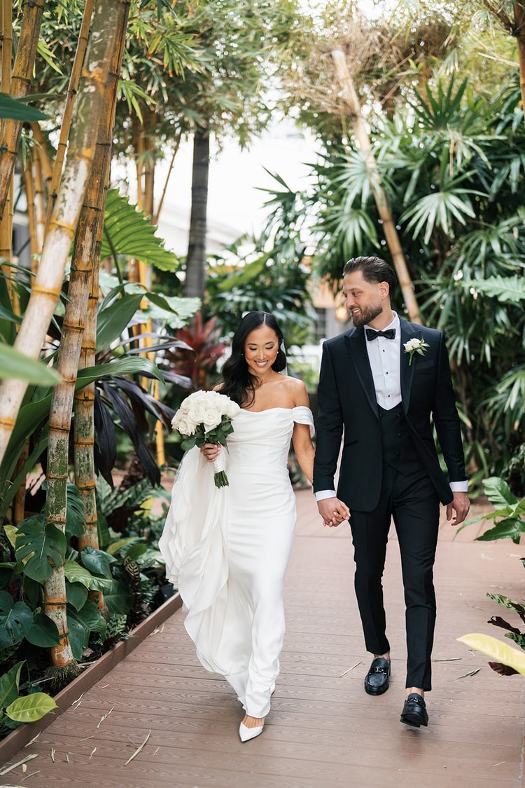 Bride and groom strolling hand in hand along a wooden path through a lush tropical garden with bamboo and broad-leafed plants, wedding photography