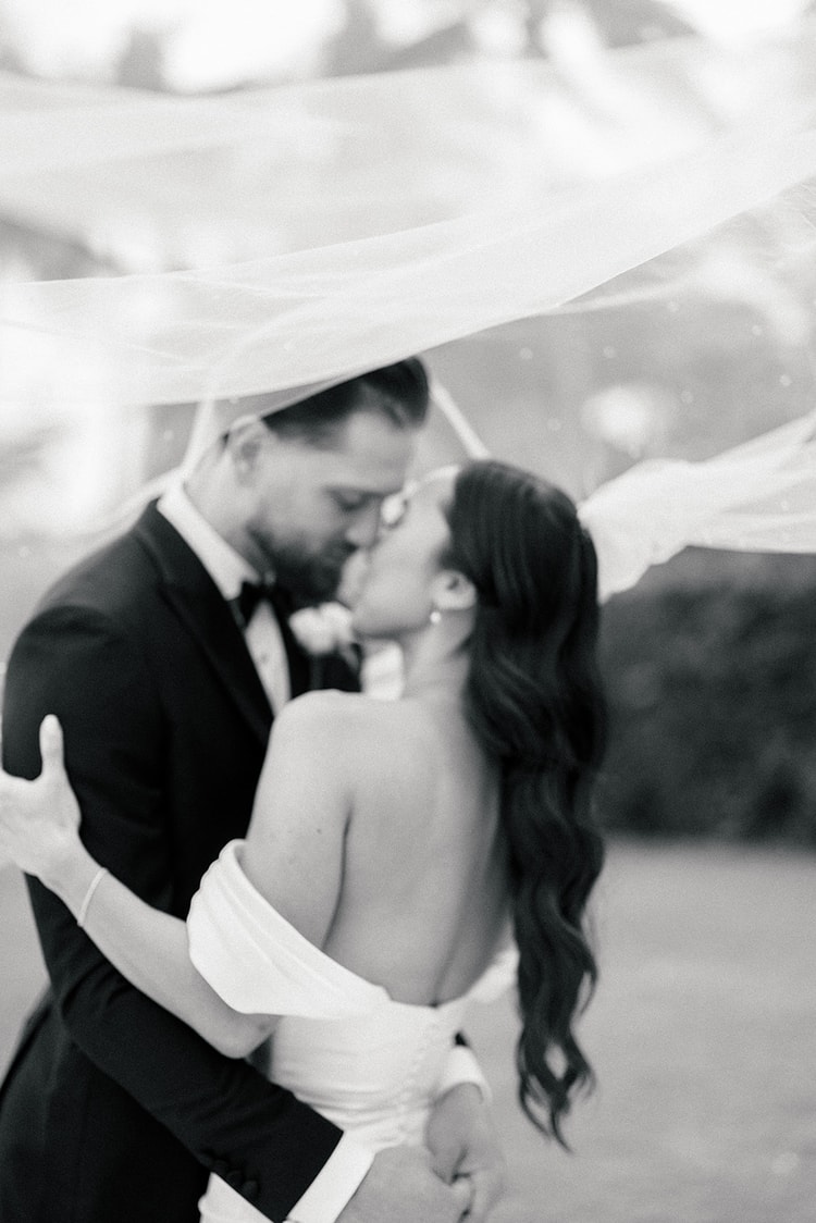 Bride and groom sharing a close kiss beneath a floating pearl-dotted veil, intimate black and white portrait, wedding photography