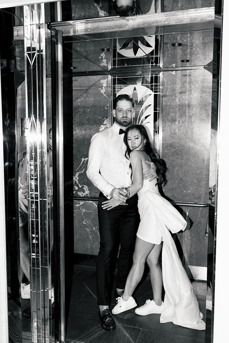 Couple in reception attire embracing inside a mirrored elevator, groom in white jacket, bride in a short white dress, black and white, wedding photography