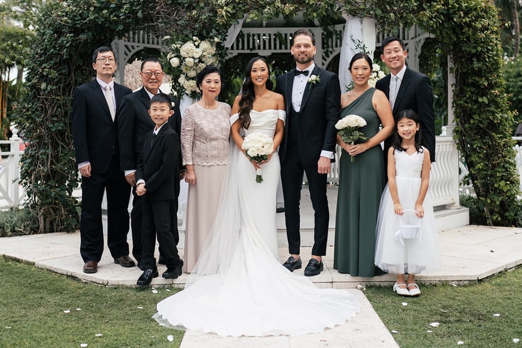 Couple with extended family posing in front of a white ivy-covered gazebo, bride holding white roses, wedding photography