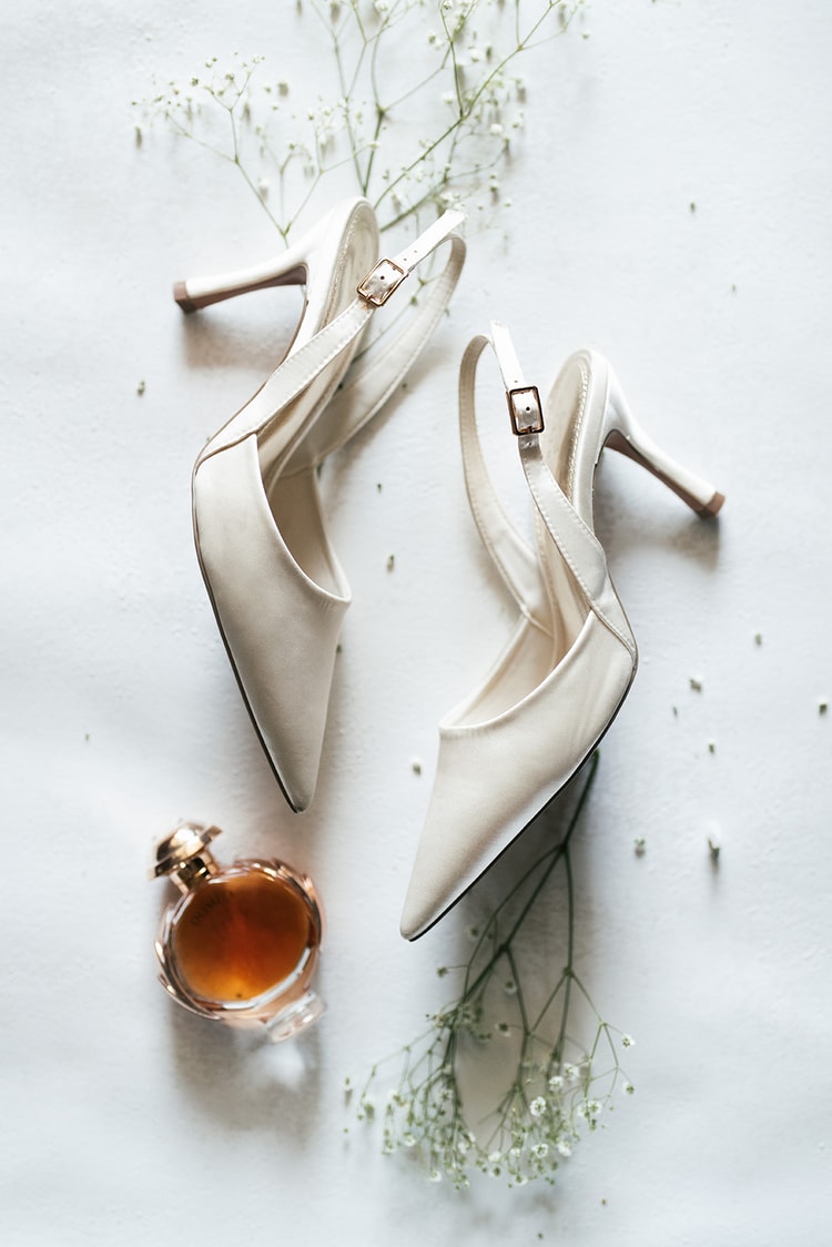 Bride's ivory slingback heels and a small perfume bottle arranged on a white surface with scattered baby's breath, getting-ready details.