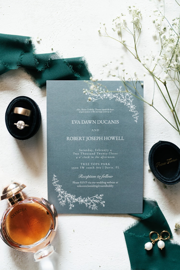 Flat-lay of the slate-blue wedding invitation suite surrounded by a perfume bottle, earrings, rings, and green ribbon details.
