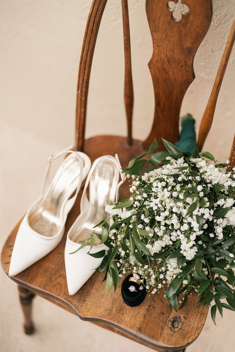 Bridal details flat-lay: ivory slingback heels, a bouquet of baby's breath and greenery, and a diamond ring in a velvet box on a wooden chair.