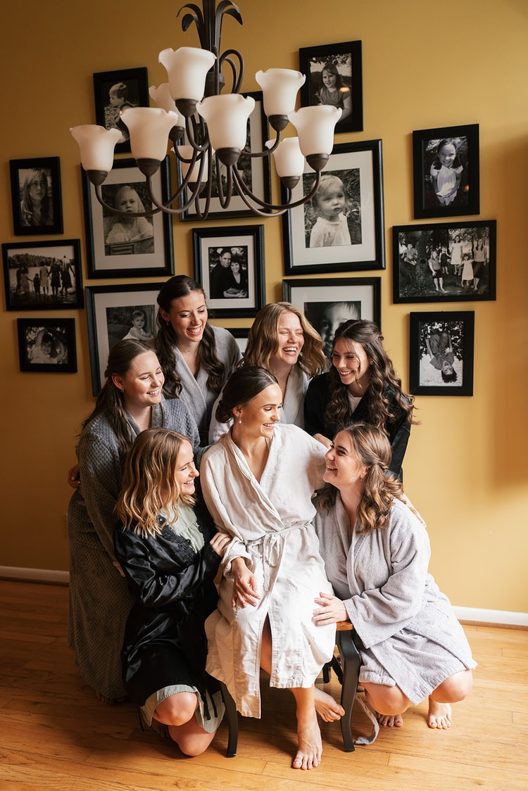 Bride surrounded by six bridesmaids in matching robes, laughing together in a yellow-walled room during getting-ready.