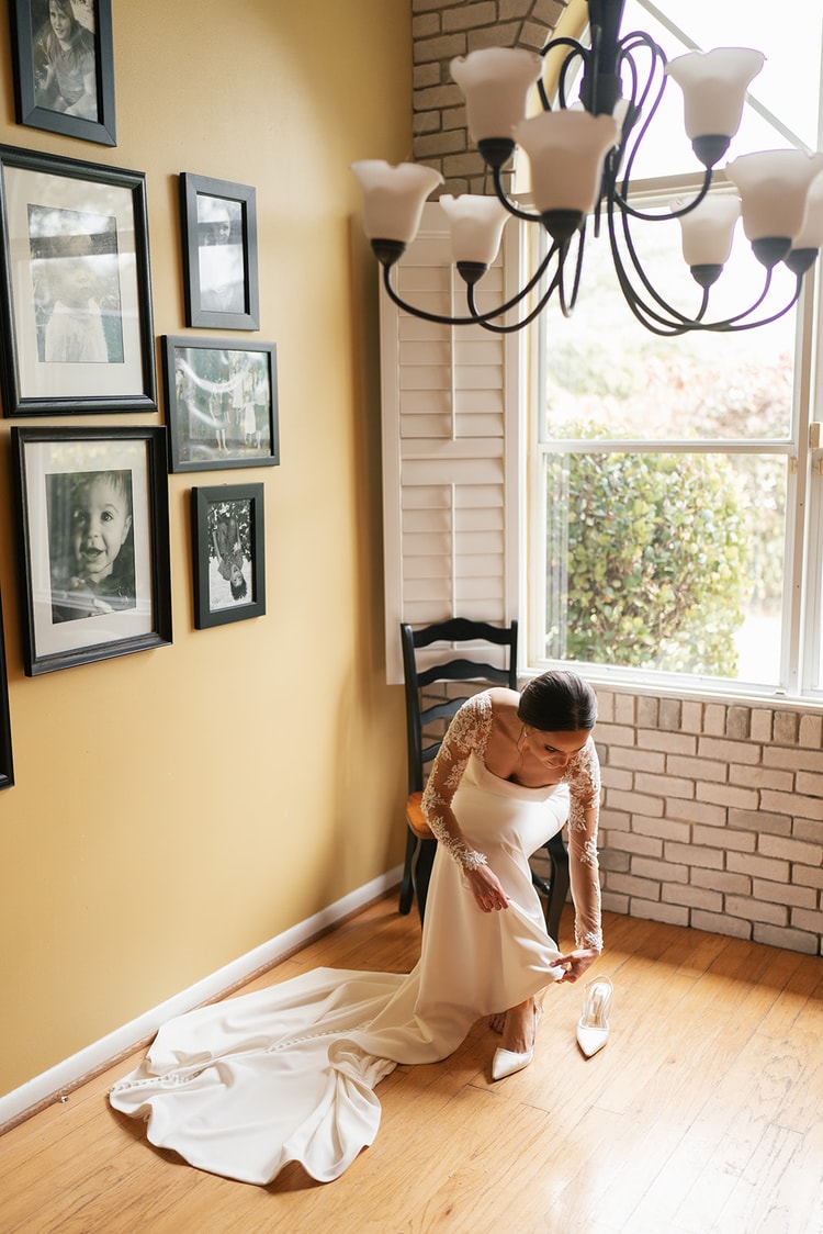 Bride in her lace-sleeved gown fastening her shoe in a warmly lit room surrounded by family portrait frames, getting ready.