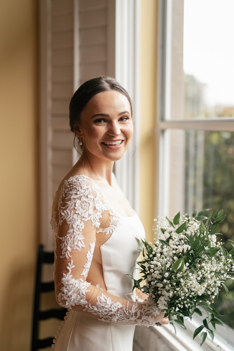 Bride smiling over her shoulder near a sunlit window, holding her greenery-and-baby's-breath bouquet, lace sleeves visible.