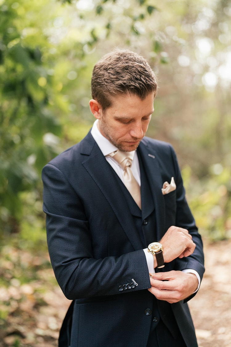 Groom in a navy three-piece suit adjusting his watch among lush garden foliage, getting-ready portrait.