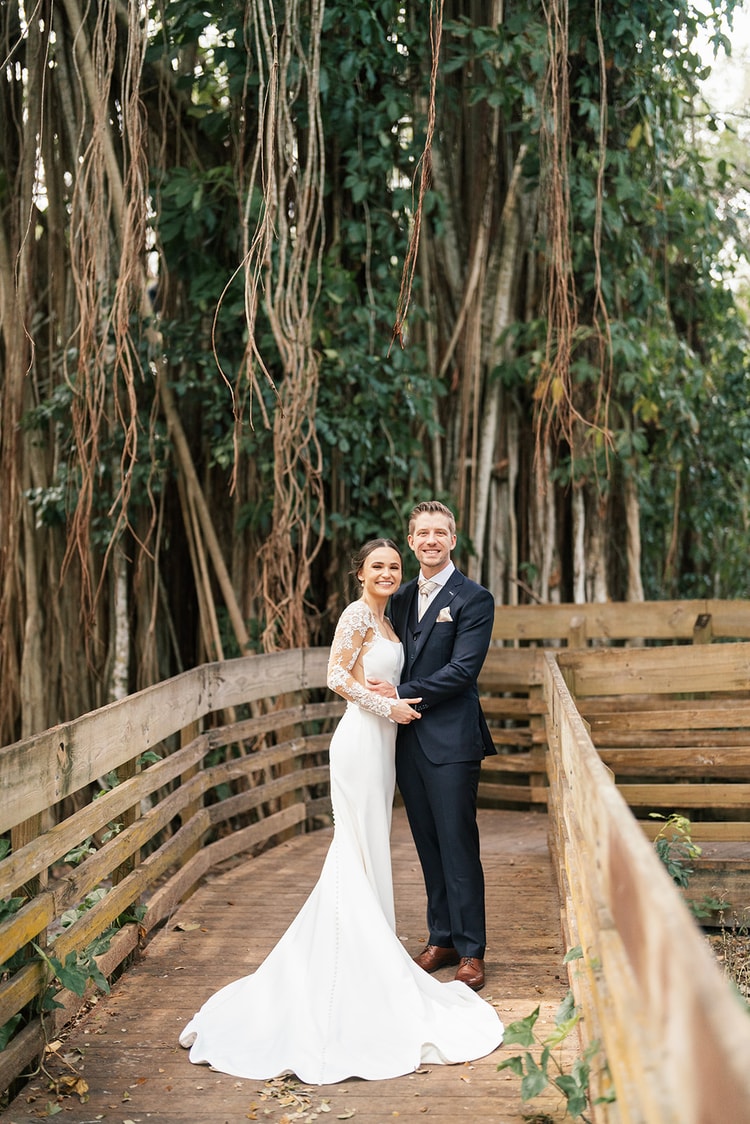 Couple posing on a weathered wooden boardwalk flanked by sweeping banyan vines, smiling at the camera.