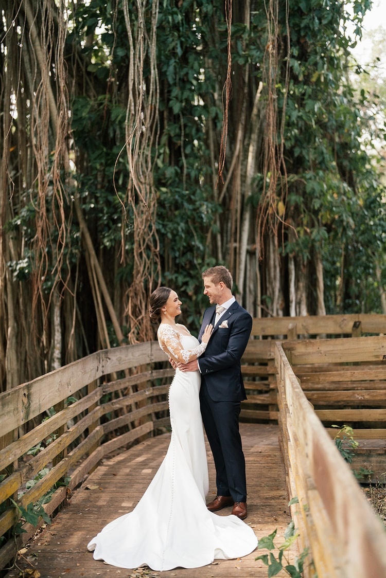 Bride and groom face each other on a rustic boardwalk surrounded by hanging banyan roots, sharing a quiet moment.