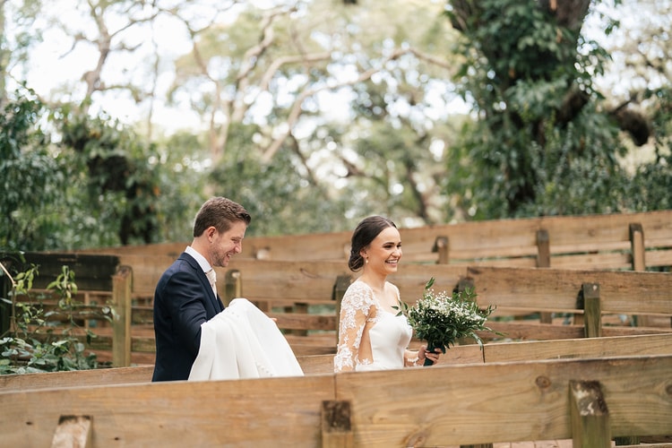 Couple walks through the wooden ceremony benches at an outdoor park venue, both laughing as the groom carries the bride's train.