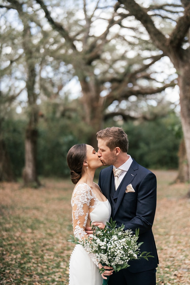 Bride and groom share a first kiss portrait under ancient oak trees, bouquet in hand, fallen leaves on the ground.