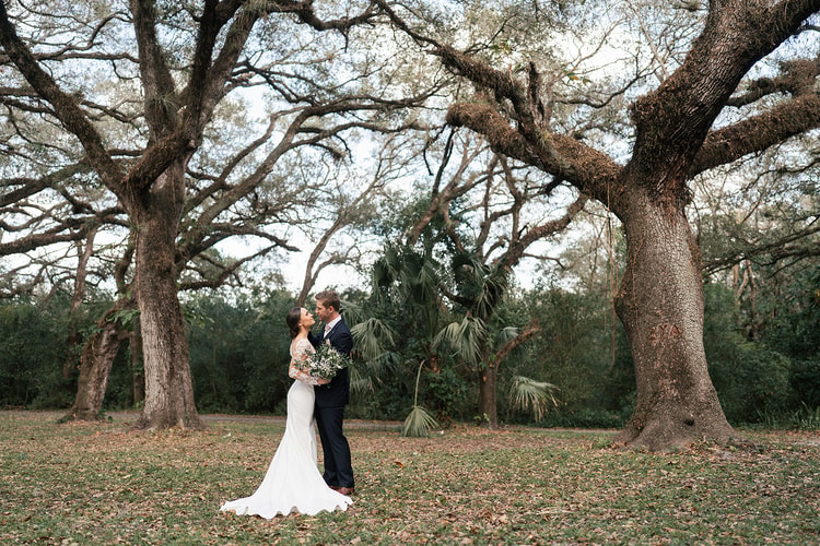 Bride and groom stand close beneath large live oaks at the edge of a clearing, a quiet portrait after the ceremony.