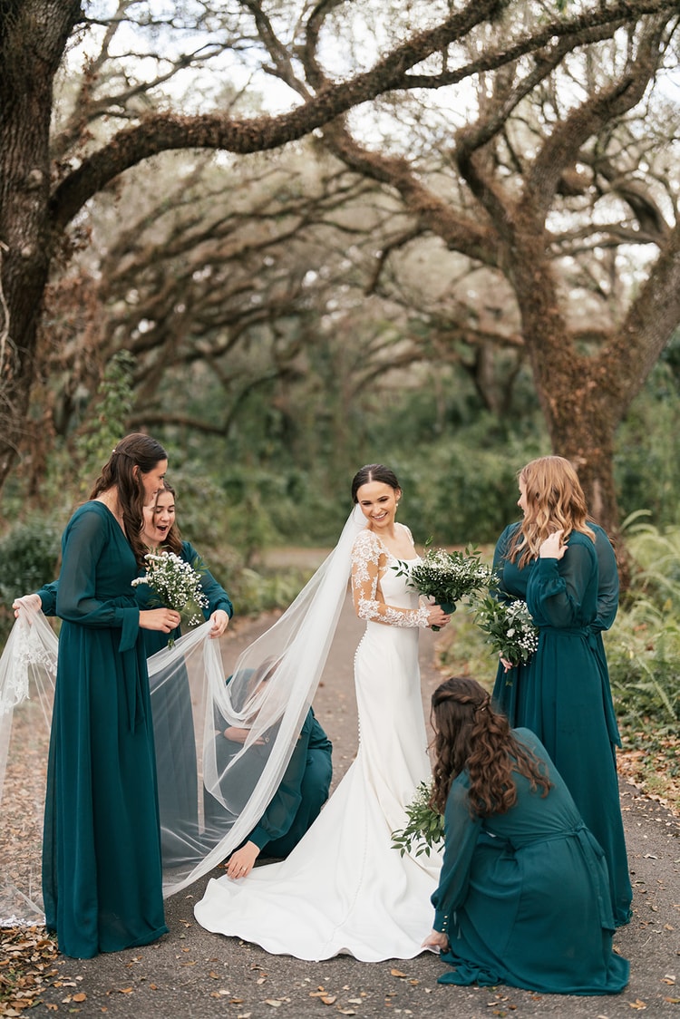 Bridesmaids in floor-length teal gowns help the bride fluff and arrange her cathedral veil under moss-draped oak trees.